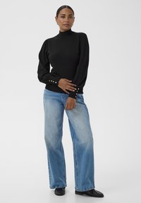 O femeie stă îmbrăcată cu un pulover negru cu mâneci bufante și guler înalt, cu butoane aurii și blugi evazați de culoare albastră deschisă, purtând loafers negri.