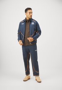 Mornarska trenirka z oranžnimi detajli, športnega videza. Vključuje jakno na zadrgo in usklajene hlače. Nosi bež vyžvanjane superge.