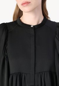 Blusa preta com um decote alto, mangas pregueadas e um fecho central com botão adornado por uma gema decorativa. Tecido de textura suave.