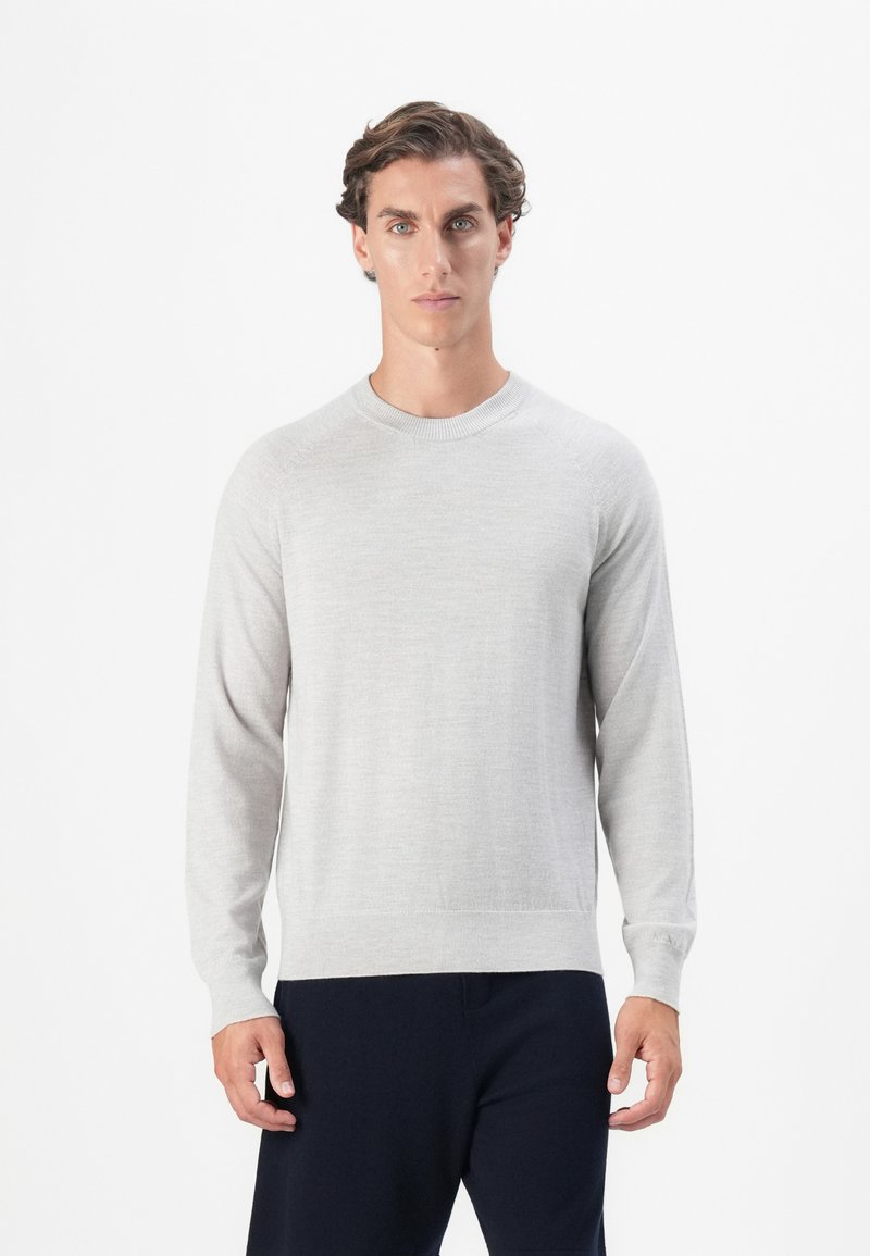 Uomo con capelli ricci corti che indossa un maglione a maniche lunghe grigio chiaro e pantaloni scuri, in piedi di fronte a uno sfondo bianco semplice.