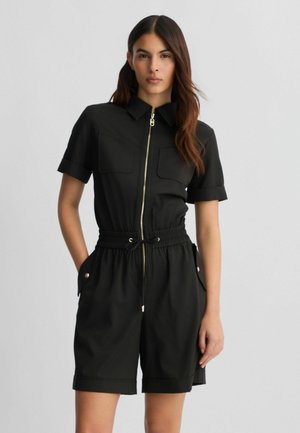 Jumpsuit court noir avec zipper sur le devant, col, taille élastique avec cordon de serrage, et deux poches avant avec accents dorés.