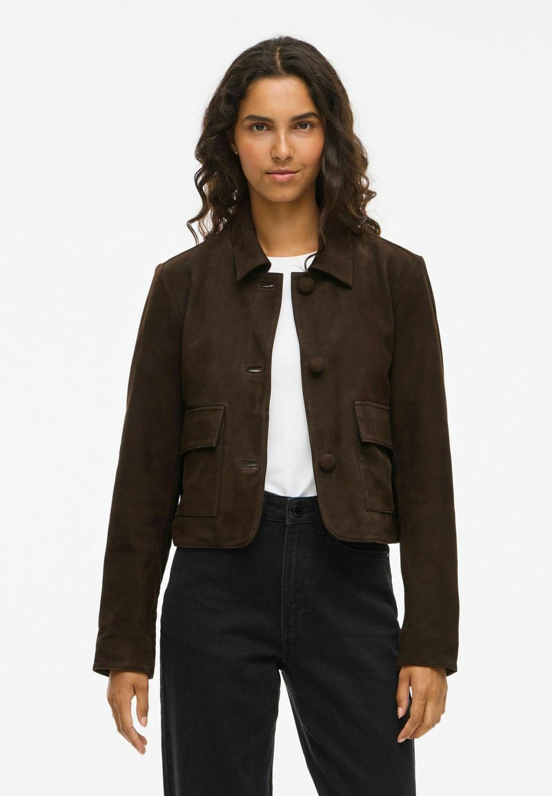 Brun ruskinds cropped jakke med krave, to forlommer og knaplukninger. Båret over en hvid skjorte, parret med sorte bukser.