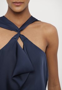 Tmavě modrý halter top s uzlem na výstřihu a zavěšenou látkou. Jemná, hladká textura; má otevřený design hrudníku a minimální prošívání.