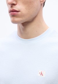 Νέος άνδρας που φοράει ένα ανοιχτό μπλε πουκάμισο με υφή και ένα μικρό λογότυπο Calvin Klein κοντά στο στήθος, σε λευκό απλό φόντο.