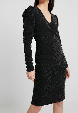 Woman wearing a fitted, long-sleeve black dress with subtle glitter, a V-neckline, ruched waist, and slightly puffed shoulders.