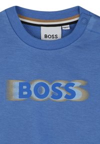 T-shirt in cotone azzurro chiaro con un grande logo stilizzato "BOSS" in blu e giallo, con collo a giro e dettaglio dei bottoni a pressione.