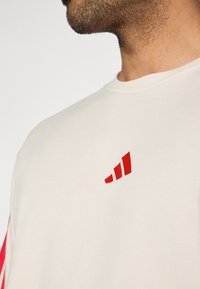 Κρεμ φούτερ με στρογγυλή λαιμόκοψη, μαλακή υφή βαμβακιού και έντονο κόκκινο αφηρημένο λογότυπο στο αριστερό στήθος.