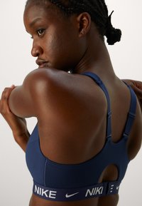 Sutien sportiv bleumarin cu bretele ajustabile, având o bandă elastică lată inscriptionată cu "Nike" și o decupare în formă de inel la spate. Textură netedă.