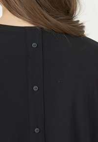 Svart t-shirt med en slät textur, som har en knäppt bakstängning med sex svarta knappar som är vertikalt placerade längs mitten.