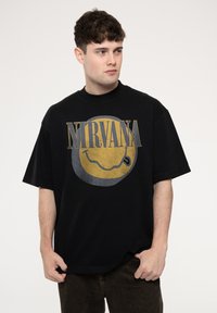 Black t-shirt with oversized fit featuring a distressed yellow smiley face graphic and "NIRVANA" text in gray, made of cotton fabric.