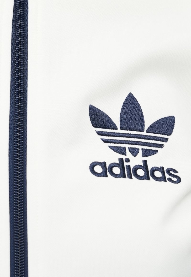 White zip-up jacket with navy blue embroidered Adidas logo and three stripes design. Smooth texture, synthetic material.