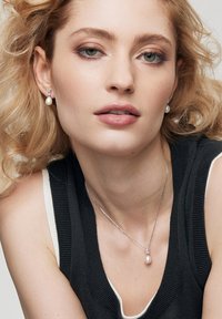 Srebrny naszyjnik z zawieszką i pasujące kolczyki z okrągłą białą perłą, noszone z czarno-białą prążkowaną bluzką. Miękkie, falujące blond włosy.