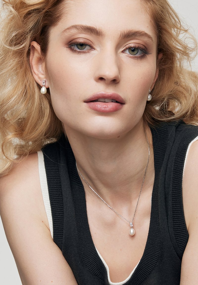 Srebrny naszyjnik z zawieszką i pasujące kolczyki z okrągłą białą perłą, noszone z czarno-białą prążkowaną bluzką. Miękkie, falujące blond włosy.