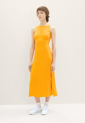 Ärmelloses, leuchtend oranges Kleid mit einem seitlichen Schlitz, aus glattem Stoff gefertigt. Kombiniert mit weißen Sneakers und hellrosa Kniestrümpfen.