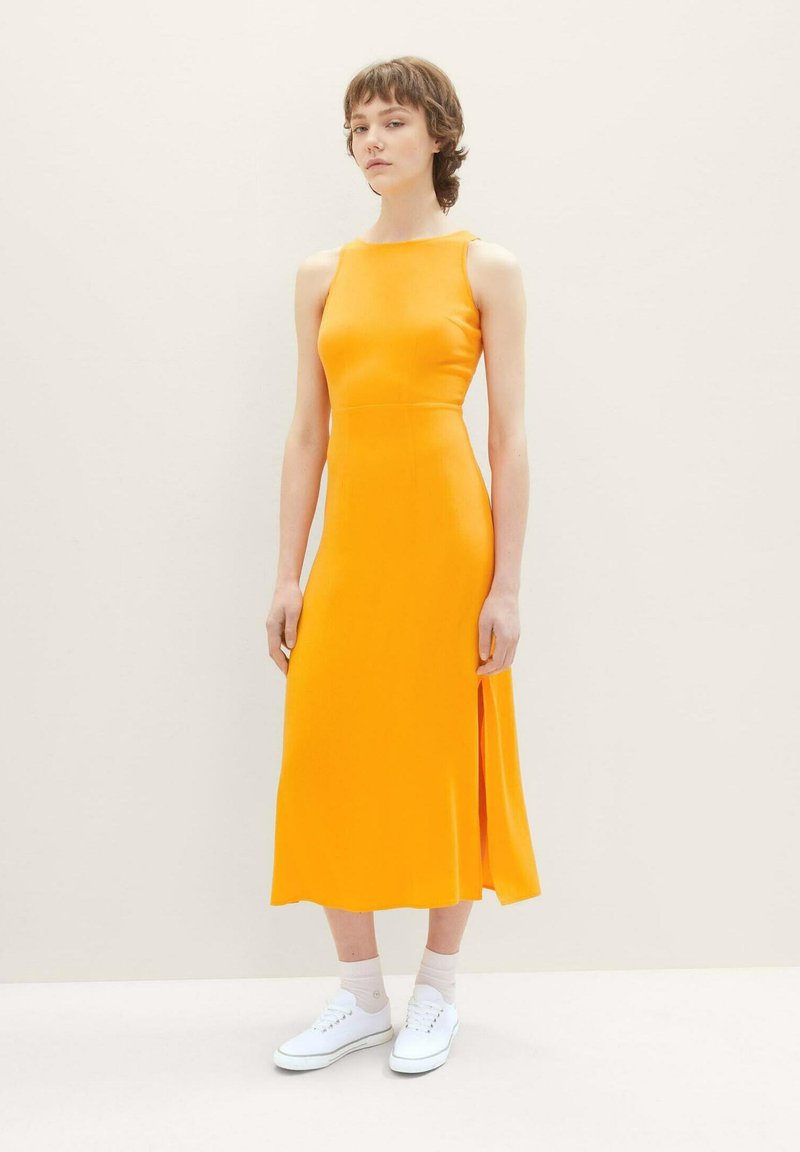 Ärmelloses, leuchtend oranges Kleid mit einem seitlichen Schlitz, aus glattem Stoff gefertigt. Kombiniert mit weißen Sneakers und hellrosa Kniestrümpfen.