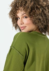 Jersey verde oliva con un corte relajado, detalles de costura visibles y una textura suave. Modelo mostrado de espaldas, con el cabello estilizado en rizos.
