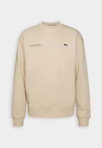 Sudadera beige de tela suave, con cuello redondo, puños acanalados y un pequeño logo con texto en el pecho.