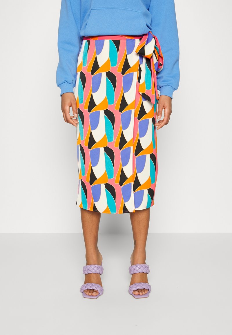 Jupe midi portefeuille à motif géométrique coloré, associée à un sweat bleu et des sandales à talons avec brides tressées lavande sur une personne debout.