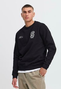 Hanorac negru din bumbac cu guler rotund. Caracteristici include un logo brodat cu "S" și detalii textile pe partea stângă a pieptului. Purtați-l cu pantaloni din catifea.