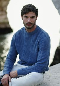 Maglione azzurro lavorato a maglia con un motivo a treccia testurizzato, maniche lunghe e scollatura rotonda, abbinato a pantaloni bianchi, ambientato in uno sfondo naturale.