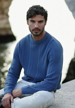 Maglione azzurro lavorato a maglia con un motivo a treccia testurizzato, maniche lunghe e scollatura rotonda, abbinato a pantaloni bianchi, ambientato in uno sfondo naturale.
