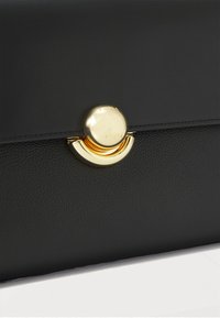 Sac en cuir noir avec un fermoir rond en or et un accent en croissant en bas. La texture est lisse avec un grain subtil.