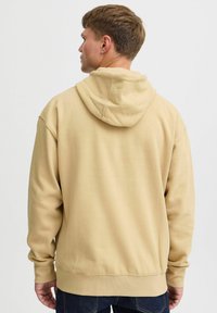 Sudadera con capucha beige de corte relajado, puños y dobladillo acanalados, y textura suave. Cuenta con un bolsillo tipo canguro en la parte frontal.