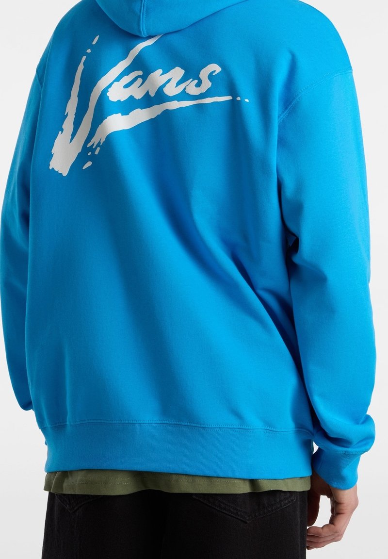 Personne portant un sweat à capuche bleu vif avec un logo « Vans » blanc stylisé sur le haut du dos, superposé à une chemise vert olive et un pantalon noir.