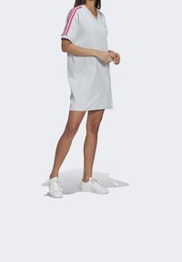 Witte sportieve jurk met een V-hals, korte mouwen en roze drie-strips accenten op de schouders, gestyled met witte sneakers.