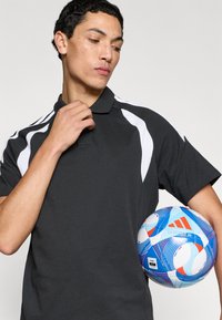 Μαύρο πόλο μπλουζάκι με λευκές λεπτομέρειες, κοντά μανίκια και υφασμάτινη υφή. Το μοντέλο κρατά μια πολύχρωμη, διακοσμημένη μπάλα ποδοσφαίρου.