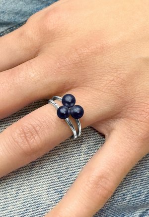 Main portant une bague argentée à double bande ornée de trois perles bleu foncé sur le majeur, reposant sur un tissu en denim.