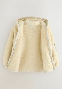 Kermanvärinen fleece-takki hupulla, jossa on pehmeä ja teksturoitu sisus, kultaiset nepparit sekä sivussa vetoketjullinen tasku.