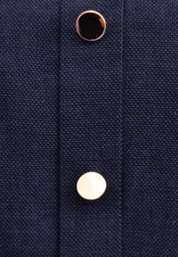 Granatowa tkanina o teksturowanym splocie z dwoma okrągłymi guzikami; jeden jest metaliczno-złoty, a drugi ma błyszczący czarny kolor.