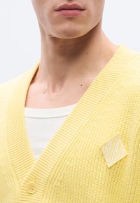 Κίτρινη πλεκτή ζακέτα με βαθύ V λαιμόκοψη, με κλείσιμο με κουμπιά και μικρό διαμαντένιο σχέδιο με ριγέ μοτίβο στο στήθος.