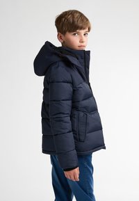 Jachetă căptușită de culoare bleumarin cu închidere cu fermoar, glugă și buzunare laterale. Prezintă secțiuni quilted și manșete ribbate pentru un plus de confort.
