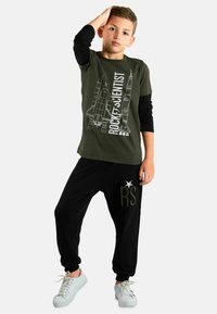 Completo per bambini composto da una maglietta verde oliva con grafica di un razzo e testo, abbinata a pantaloni neri e sneakers bianche.