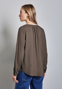 Brun långärmad blus med veckdetaljer på baksidan, rund halsringning och något lös passform, parat med blå denimjeans.