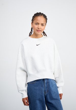 Lys grå, oversized sweatshirt med rund hals og ribbestrikket nederkant; har en liten svart Nike-logo på brystet. Kombinert med blå denimjeans.
