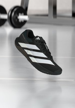 Chaussure de sport noire à rayures blanches flottant au-dessus d’un sol de salle de sport, équipement de musculation flou en arrière-plan.