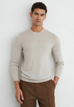Mand iført en lysebeige langærmet sweater og brune bukser, der står op ad en hvid væg med den ene hånd i lommen.