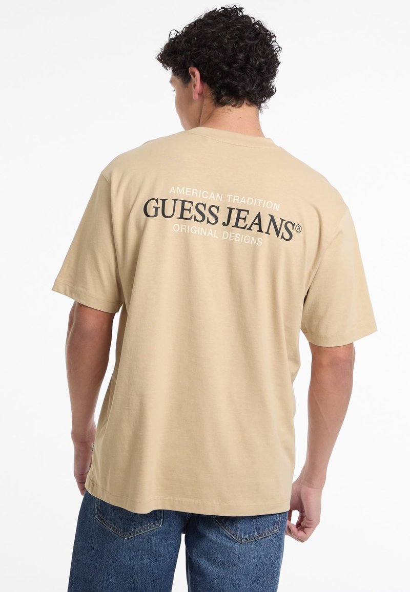 Bēšas krāsas kokvilnas t-krekls ar īsām piedurknēm, aizmugurē ar melnu tekstu: "AMERIKĀŅU TRADĪCIJA GUESS JEANS ORIĢINĀLIE DIZAINS."