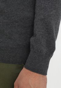 Ärmelsaum eines dunkelgrauen, gestrickten Pullovers mit gerippter Struktur. Das Material wirkt weich und warm. Eine Hand ist sichtbar, darunter ein olivgrünes Kleidungsstück.