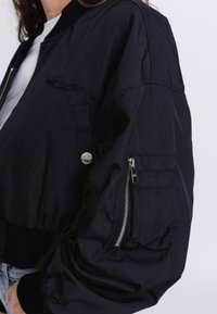 Black bomber jacket featuring a soft fabric, ribbed cuffs, and an embroidered logo. Includes a zippered pocket on the sleeve for functionality.
