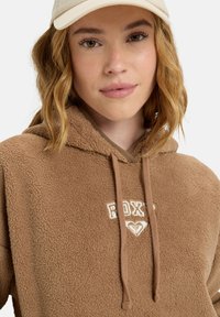 Brun fleece hættejakke med en blød tekstur, der har en kængurulomme og hvid "ROXY" logo broderi på fronten. Justerbar snor i hætten.