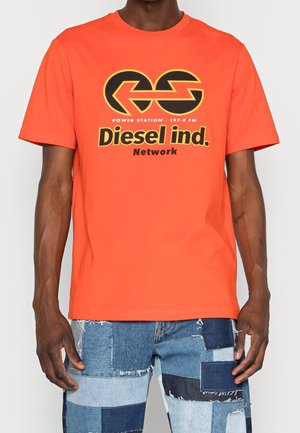 Mężczyzna ubrany w pomarańczową koszulkę Diesel ind. Network i niebieskie dżinsy patchworkowe, stojący na jednolitym jasnym tle.