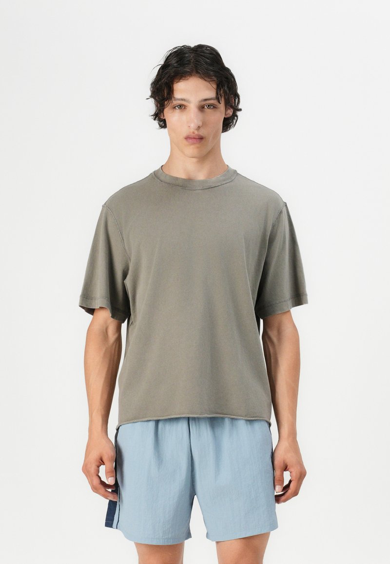 Olive green short-sleeve t-shirt with a round neck, relaxed fit, and visible stitching details, paired with light blue shorts.