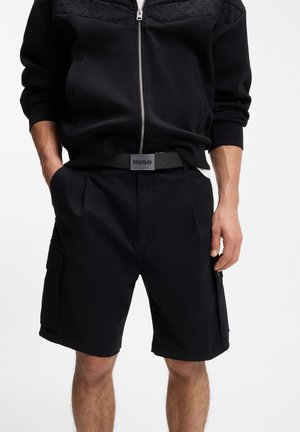 Mand iført sort lynlåstrøje og sorte cargoshorts med sort bælte med et spænde med "HUGO"-logo.