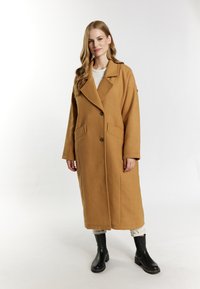 Palton lung din lână de culoare cămilă, cu guler tăiat, două buzunare frontale și sistem de închidere cu buton. Purta un top crem și pantaloni bej, asortați cu bocanci negri.