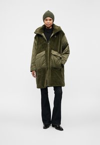 Olivgrüner, langer, mit Reißverschluss verschließbarer Mantel mit Fleece-Textur; verfügt über große frontale Taschen und einen hohen Kragen. Getragen über einem schwarzen Oberteil und dunkler Jeans.