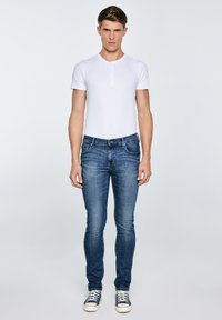 Junger Mann steht frontal, trägt ein weißes Henley mit kurzen Ärmeln, schlanke blaue Jeans und dunkle Sneaker vor einem schlichten Hintergrund.